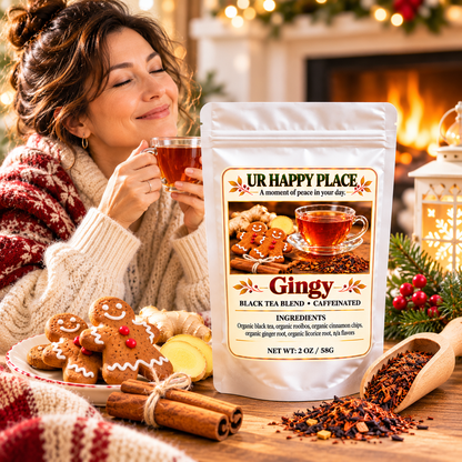 A woman in a cozy knit sweater holds a glass of ginger tea beside a stand-up pouch of UR Happy Place Ginger Black Tea Blend. The scene is styled with gingerbread cookies, cinnamon sticks, loose tea, and warm holiday decor in front of a fireplace.